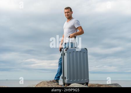 Un jeune homme en Jean et une chemise blanche se tient avec une valise grise sur le bord de mer sur d'énormes rochers contre un ciel avec des nuages et regarde dans la caméra Banque D'Images