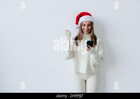 Excitée jeune Santa femme tenir utiliser le téléphone cellulaire type dire oui a découvert grand gagner faisant le geste gagnant sur fond blanc Banque D'Images