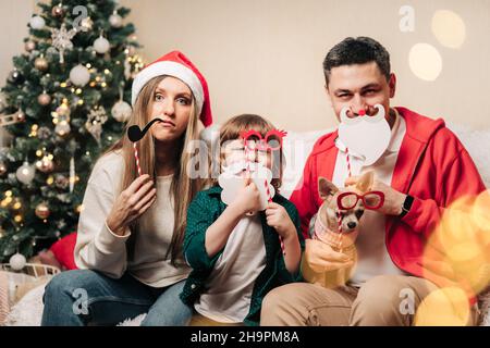 Portrait d'une famille heureuse avec un enfant, un fils et un chiot tenant des accessoires festifs pour une exposition de photos.Mère en chapeau de père Noël, père, enfant garçon et chien en chandail Banque D'Images