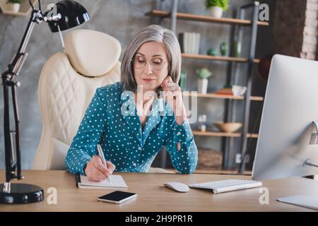 Photo sérieux calme vieux joli gris cheveux femme porter des lunettes écrire note Manager regarder à l'intérieur dans le poste de travail Banque D'Images