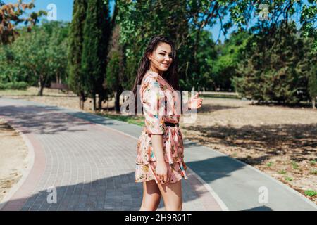 belle femme brune dans une robe avec des fleurs marche le long de la rue et le parc Banque D'Images