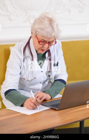 Femme d'âge moyen médecin utilisant un ordinateur portable, écrivant des notes avec le stéthoscope sur la table.Concept de soins de santé.Médecin écrivant et prenant des notes Banque D'Images