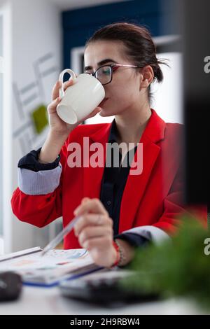 Manager en blouson rouge au bureau de démarrage en train de boire à partir d'une tasse blanche.Employé avec des lunettes ayant un café ou une pause thé au bureau.Femme d'affaires prenant une pause de la lecture de la carte sur le presse-papiers. Banque D'Images