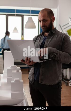 Architecte tenant un ordinateur portable comparant des plans avec la maquette de gratte-ciel dans le bureau d'architecture.Ingénieur avec ordinateur portable regardant les plans de construction de la propriété immobilière. Banque D'Images