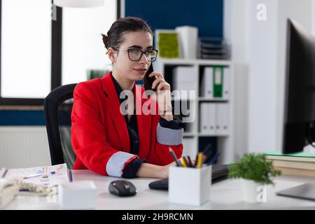 Le propriétaire d'une petite entreprise est confiant et se trouve sur le téléphone intelligent, assis au bureau.Employé avec des lunettes regardant l'écran de l'ordinateur tout en parlant au téléphone.Manager en blouson rouge dans la conversation sur téléphone portable. Banque D'Images