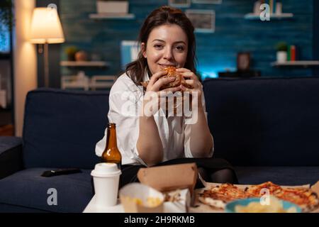 Femme souriante tenant un savoureux hamburger entre les mains et regardant la comédie télévisée sitcom sur le canapé devant la table avec menu de restauration rapide à emporter.Bonne personne regardant un spectacle télé avec un dîner à emporter. Banque D'Images