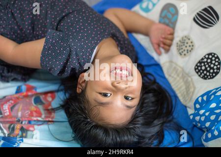 Une belle petite fille avec de longs cheveux noirs avec des dents manquantes souriant sur le lit, tirer à partir de l'angle élevé Banque D'Images