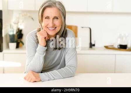 Élégante charmante femme d'âge moyen aux cheveux gris, assise sur le comptoir dans une cuisine moderne et lumineuse, regarde l'appareil photo, souriant, met son menton sur les mains.Concept de bien-être Banque D'Images