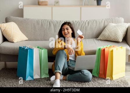 Femme arabe enthousiaste se sentant heureuse de la grande vente, en utilisant un ordinateur portable et montrant la carte de crédit, assis sur le sol avec des sacs-cadeaux Banque D'Images