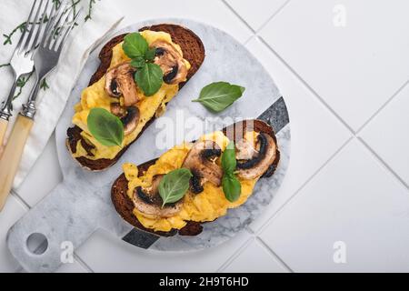 Œufs brouillés aux champignons frits et basilic sur du pain sur fond de table blanc.Petit-déjeuner ou brunch maison.Œufs brouillés et champignons Banque D'Images