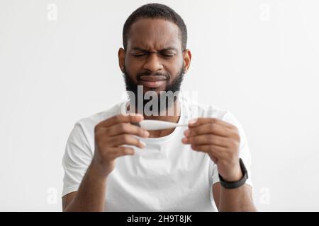 Portrait d'un homme barbu noir millénaire mécontent regarde un thermomètre à haute température sur fond de mur blanc Banque D'Images