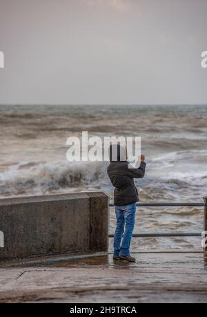 jeune femme utilisant un téléphone portable smartphone pour photographier les vagues qui se brisent lors d'une tempête d'hiver sur la côte de l'île de wight, les vagues qui s'écrasont mur Banque D'Images