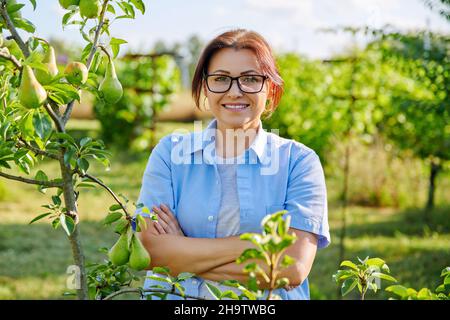 Portrait d'une femme d'âge moyen souriante et confiante avec bras croisés dans un verger, près d'un poirier Banque D'Images