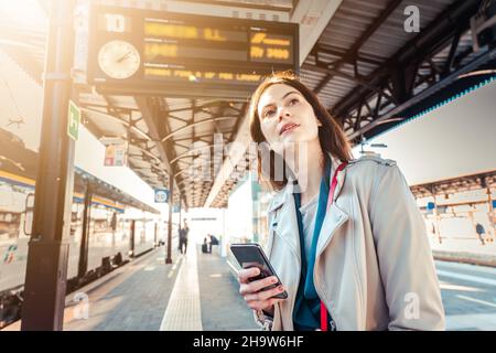 Une jeune femme avec des heures de départ derrière elle attendant son train tout en tenant son téléphone mobile - une femme regardant l'horloge dans la gare Banque D'Images