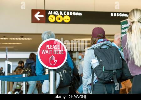 Les voyageurs attendent la file d'attente à un aéroport très fréquenté pendant une période de covid à côté d'un panneau Please WAIT here. Banque D'Images