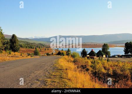 Une route de campagne descend de la colline vers des maisons sur les rives d'un lac pittoresque surplombant les montagnes enneigées.Plateau d'Ulaganskoe, Altaï, si Banque D'Images