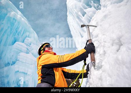 Une femme escalade une chute d'eau gelée dans un casque avec une hache de glace dans une veste orange dans les montagnes.Sport alpinisme et alpinisme concept. Banque D'Images