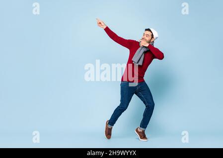 Jeune heureux beau homme caucasien portant une tenue de Noël dansant et pointant les mains vers le haut dans un arrière-plan isolé bleu clair Banque D'Images