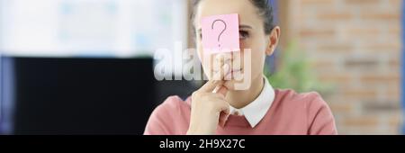 Jeune femme pensive assise à la table avec un autocollant avec un point d'interrogation sur son front Banque D'Images