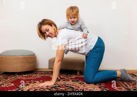 Une mère heureuse et son petit enfant jouent ensemble à la maison.Une mère conduit sa fille sur son dos.Jeux familiaux. Banque D'Images