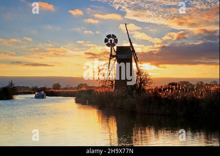 Un bateau sur la rivière Ant sur les Norfolk Broads approchant le moulin de drainage de la Fen de Turf avec un coucher de soleil rétroéclairé près de Ludham, Norfolk, Angleterre, Royaume-Uni. Banque D'Images