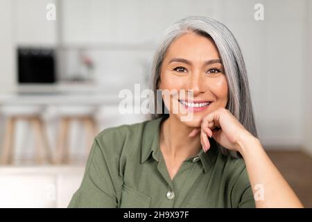 Gros plan sur une femme âgée aux cheveux gris à l'intérieur de la maison, charmante femme asiatique mûre regardant l'appareil photo et souriant, menton reposant à portée de main, femme sereine passe un week-end à la maison Banque D'Images