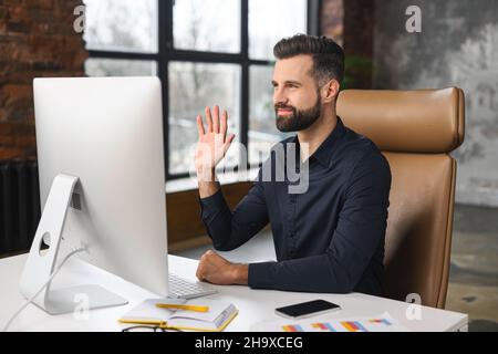 Un employé de bureau utilisant une application informatique pour la connexion vidéo avec des collègues à distance, assis devant le PC et en agitant dans une webcam, un jeune responsable réussi a impliqué une réunion virtuelle, un appel vidéo Banque D'Images