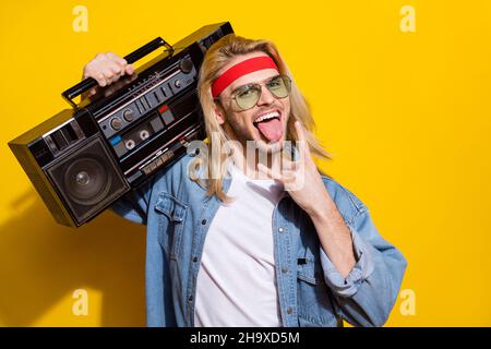 Photo de charmant mec impoli vêtu d'une chemise en denim souriant montrant la langue rock signe tenant la boîte de perche isolé couleur jaune fond Banque D'Images