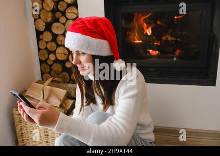 Gros plan de la jeune femme souriante fêtez Noël à la maison message texte message d'accueil sur le téléphone portable.Les femmes du millénaire heureux ont un appel téléphonique près de la cheminée Banque D'Images