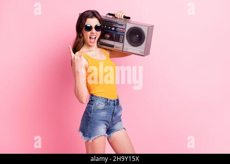 Photo d'une dame impolie vêtue jaune tank-top verres foncés tenant la boîte de perche montrant le signe de roche dure isolé couleur rose arrière-plan Banque D'Images