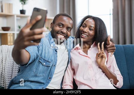 Beau et heureux couple assis dans des câlins sur un canapé confortable et de prendre selfie sur un smartphone.Homme et femme africains positifs utilisant des gadgets modernes pendant le temps libre à la maison. Banque D'Images