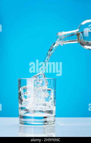 De l'eau fraîche coule de la bouteille dans le verre avec des glaçons sur fond bleu Banque D'Images