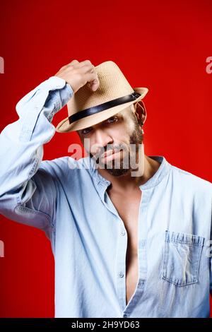 Homme cubain ou latino en chapeau et chemise sur fond rouge.Studio portrait d'un homme barbu de 30-40 ans.Émotion neutre.L'homme prend son chapeau de la tête. Banque D'Images