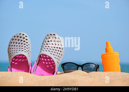 Gros plan sur des chaussures de sabots, de la crème solaire et des lunettes de soleil noires de protection sur une plage de sable au bord de la mer tropicale par temps chaud et ensoleillé. Concept de vacances d'été. Banque D'Images