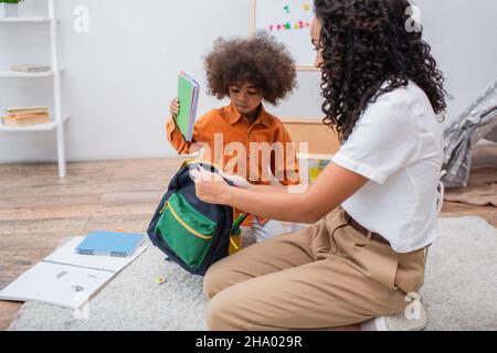 Enfant afro-américain tenant le carnet près de la mère avec sac à dos dans la salle de séjour Banque D'Images