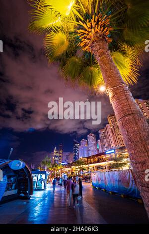 Les touristes se promènent le long de la promenade bordée de palmiers à côté de la plage dans le quartier de la Marina de Dubaï, Dubaï, Émirats Arabes Unis, avec des gratte-ciels qui s'élèvent en arrière-plan Banque D'Images