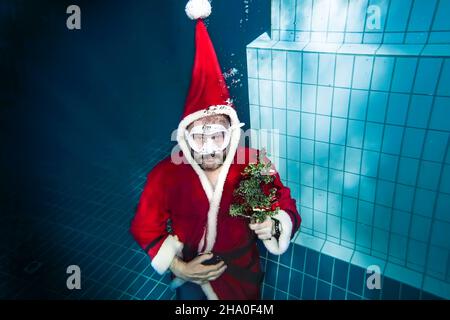 Un homme en costume de Père Noël et un masque blanc tient un arbre de Noël décoré sous l'eau dans une piscine profonde.Le Père Noël sous-marin Banque D'Images