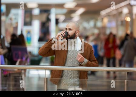 Un homme boit du café tout en prenant un masque médical dans le centre commercial. Un homme chauve garde la distance sociale. Banque D'Images