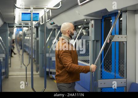 Un homme avec une barbe dans un masque médical pour éviter la propagation du coronavirus se prépare à quitter la voiture de métro tenant la main courante. Un homme chauve Banque D'Images