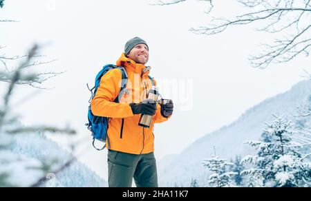 Smiling Man under Snowfall habillé blouson Softshell orange vif avec une thermos boisson chaude en trekking route des montagnes d'hiver. Peop actif Banque D'Images