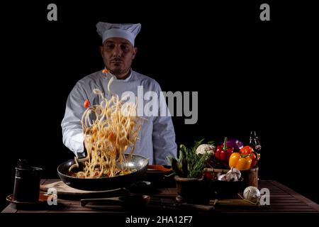 Chef masculin en uniforme blanc tenant une poêle à frire, sautant des spaghetti avec des légumes frais volant dans l'air avant de servir dans un restaurant Banque D'Images