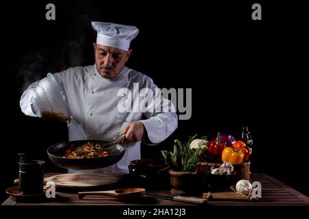 Chef masculin en uniforme blanc versant des spaghetti bouillis dans le wok pour cuire des pâtes avec des légumes.Arrière-plan de la préparation d'un plat italien traditionnel Banque D'Images