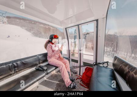 Femme skieuse utilisant l'application de téléphone dans la télécabine de ski pendant les vacances de ski.Fille souriant en utilisant un smartphone mobile portant des vêtements de ski, un casque et des lunettes Banque D'Images