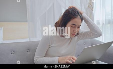 Jeune femme stressée tenant la tête dans les mains et regardant l'écran d'ordinateur portable, ayant des problèmes avec le projet, assis sur le canapé dans le salon. Banque D'Images