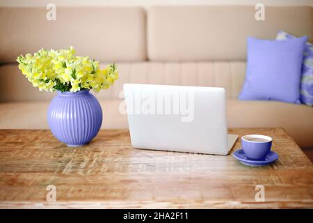 Ordinateur portable ouvert, tasse de café et vase très péri avec de belles fleurs jaunes sur table en bois rétro texturé dans un salon minimaliste avec canapé beige Banque D'Images