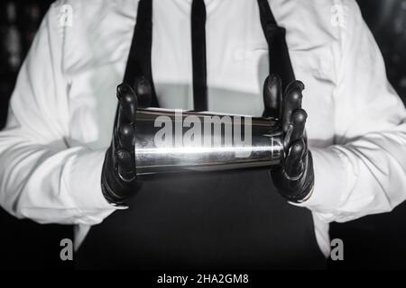 Les mains d'un barman professionnel en gants de caoutchouc latex tiennent un outil pour préparer et mélanger un shaker à cocktail alcoolisé, en gros plan. Banque D'Images