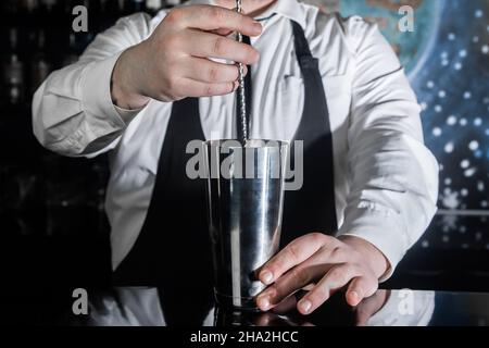 Les mains d'un barman professionnel interfèrent avec une cuillère à bar le contenu d'un shaker métallique pour préparer et mélanger des cocktails alcoolisés. Banque D'Images