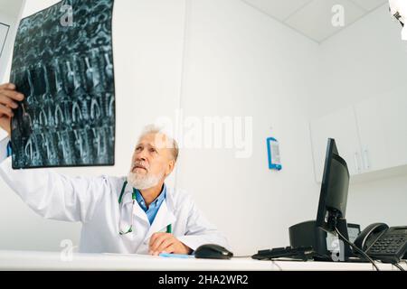 Vue à faible angle d'un médecin adulte senior sérieux qui examine l'image IRM du rachis d'un patient assis sur un bureau avec un ordinateur dans une salle de cabinet médical Banque D'Images