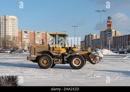 Une chargeuse sur pneus avant élimine la neige d'une place de la ville après une chute de neige sur fond de bâtiments résidentiels par temps ensoleillé. Banque D'Images