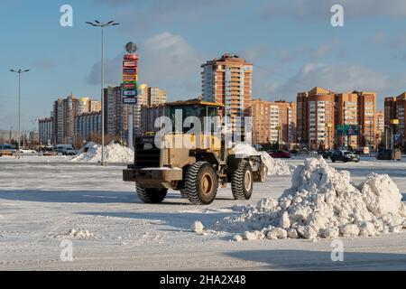 Une chargeuse à godets élimine la neige des déneigements sur une place de la ville après une chute de neige sur fond de bâtiments résidentiels par une journée ensoleillée. Banque D'Images